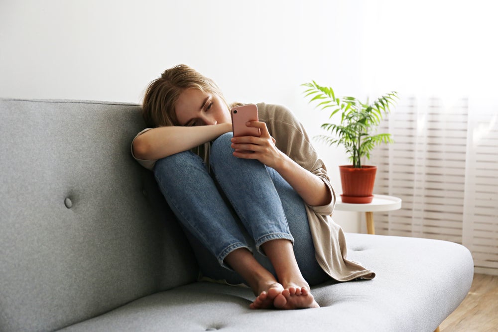 Young lady sitting on the couch with cellphone in her hand. She looks anxious about something with her head half buried in her arm.