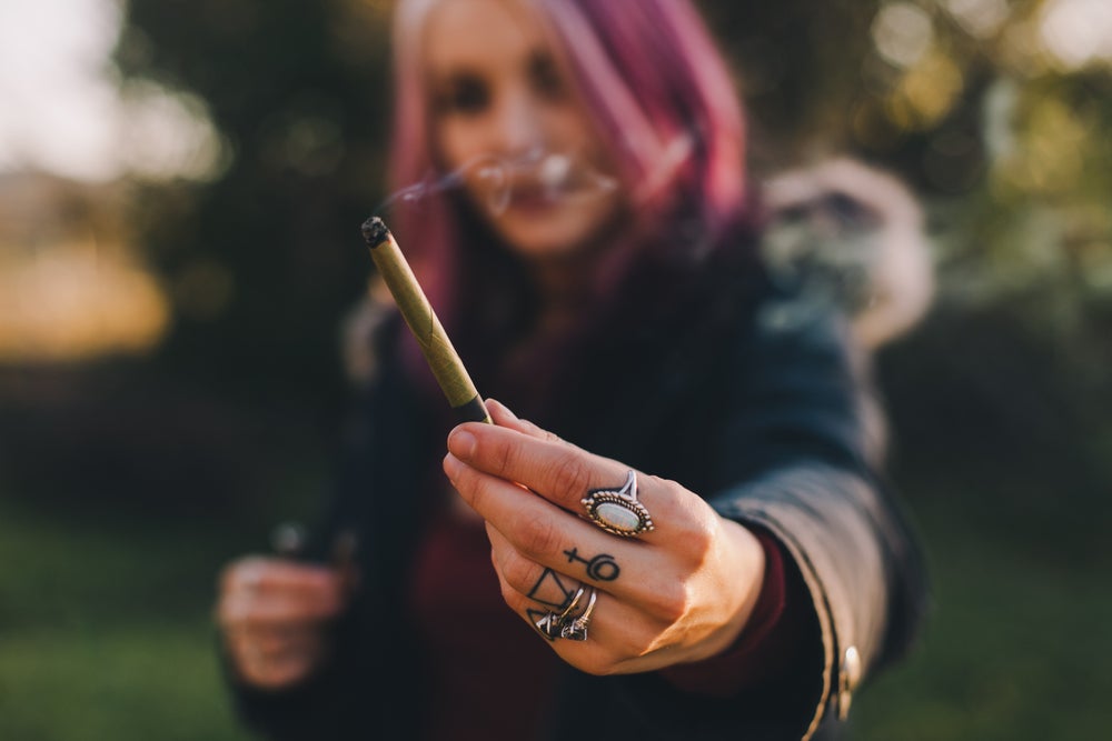 Young lady with pink hair holding out a joint with her left hand.