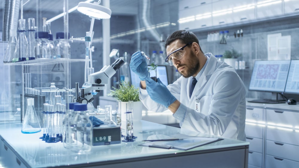 Scientist with vial containing soil and weed bud. He is holding a syringe with red liquid in his other hand.